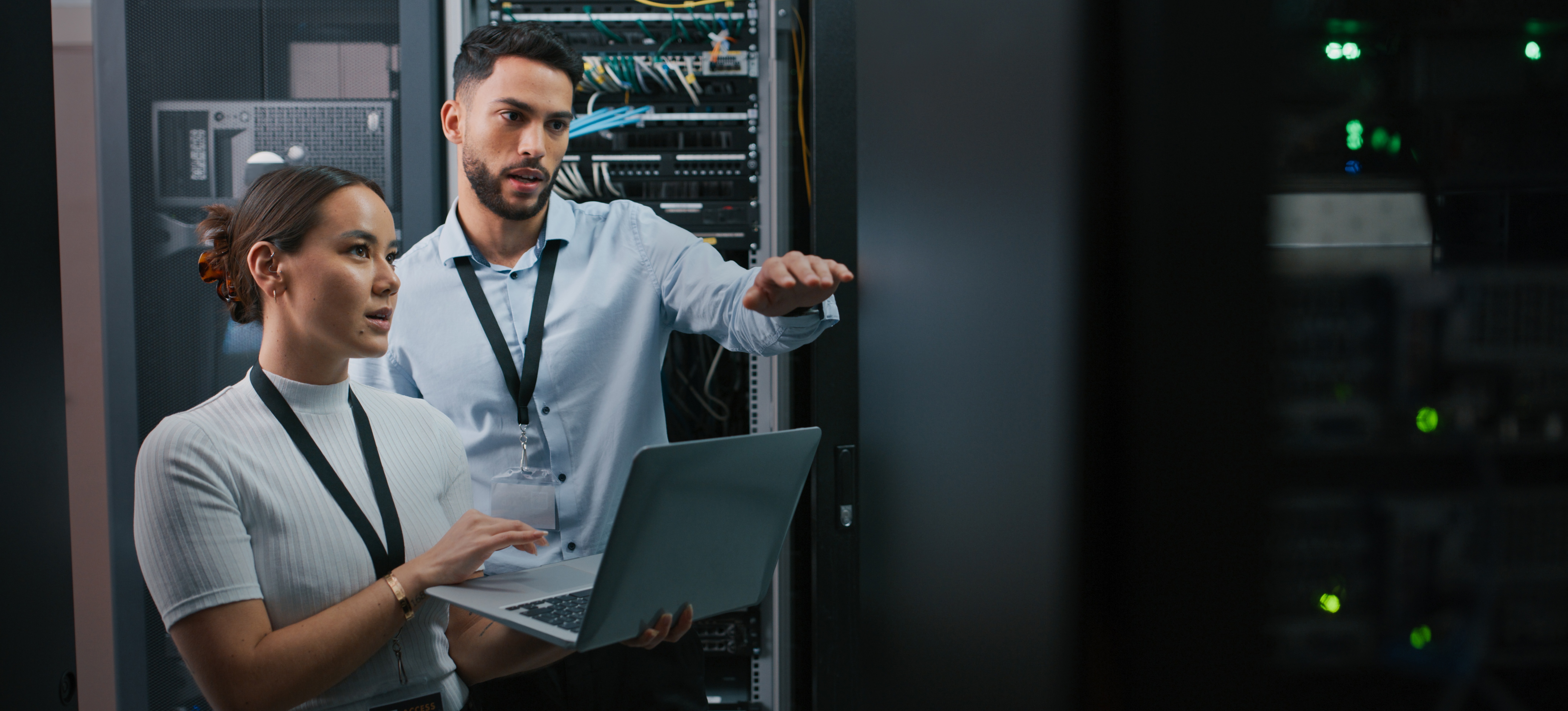 [Featured Image] Two computer engineers stand in a server room and discuss strategies.
