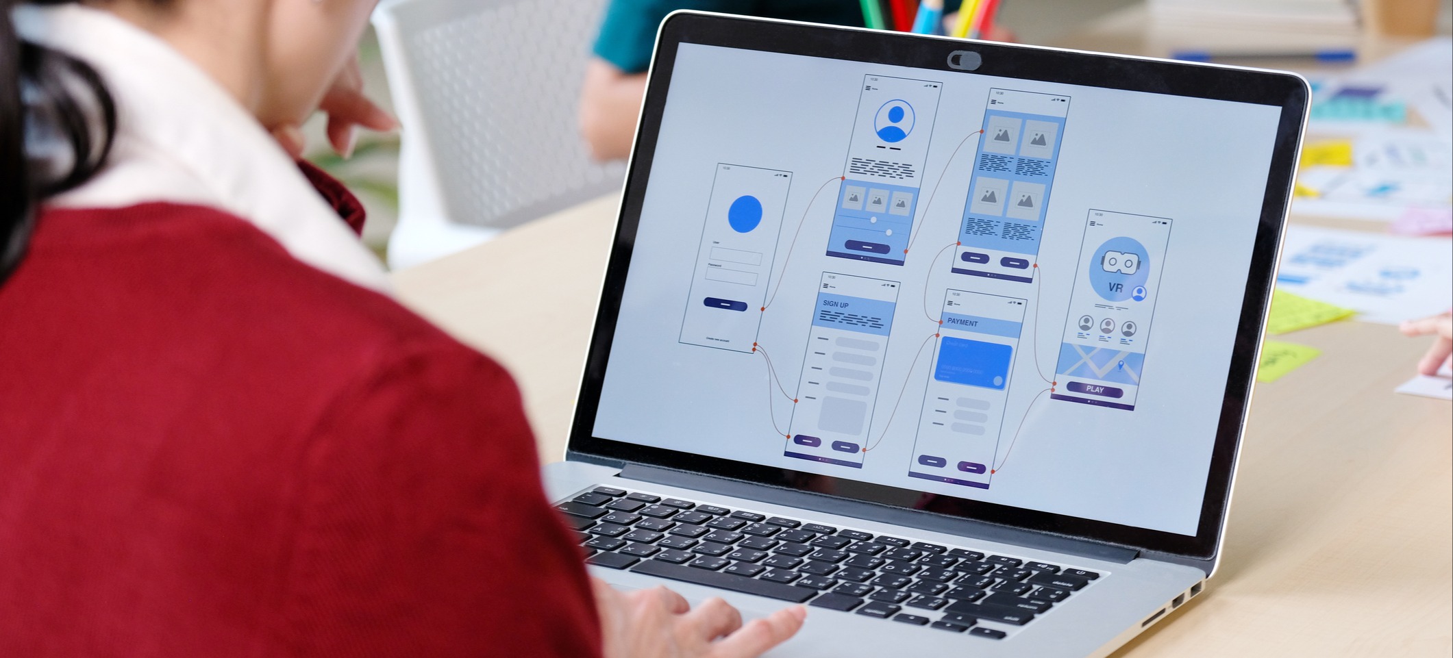 [Hauptbild] Ein UX-Designer in einem roten Pullover arbeitet an einem gemeinsam genutzten Schreibtisch an seinem Laptop an Wireframes für einen High-Fidelity-Prototyp.