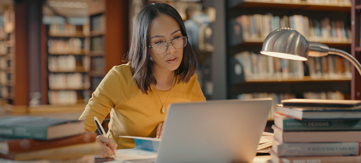 [Featured Image] A lawyer conducts research on a laptop and with books while sitting at a table in a library and writing on a notepad.
