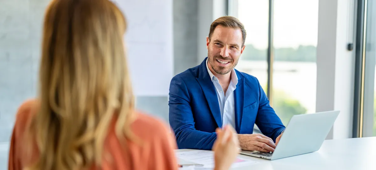 [Featured Image] An interviewer sits at a desk with a laptop, reviewing a candidate's computer engineering resume while smiling and engaging in a conversation during a computer engineering job interview.
