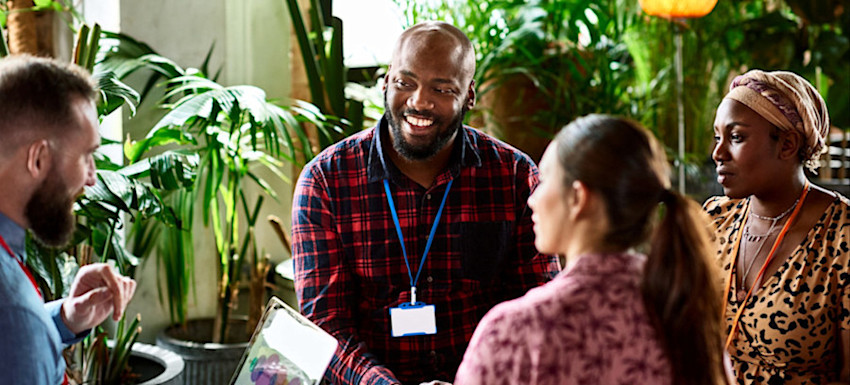[Featured Image] A project management team sits in an informal office setting with many indoor plants and discusses their current project while one holds a laptop displaying charts.
