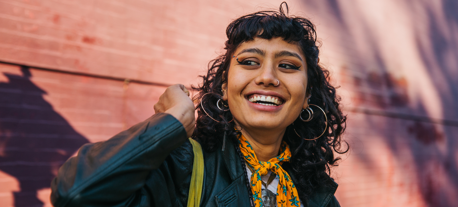 [Featured image] A young person with curly black hair and big hoop earrings, wearing a black jacket and a color scarf, smiles at someone off-camera. 
