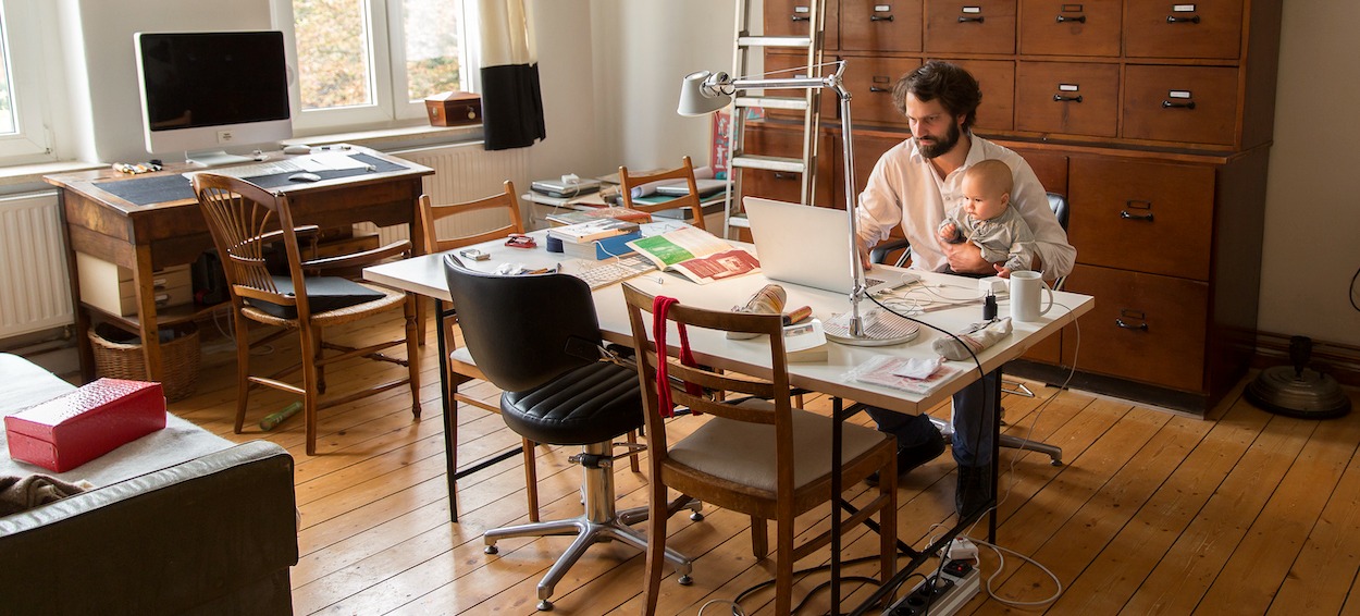 [Featured image] A UX designer is working from home holding a baby as they type on their laptop.