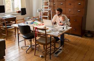 [Featured image] Employee working from home holding a baby as they types on their laptop