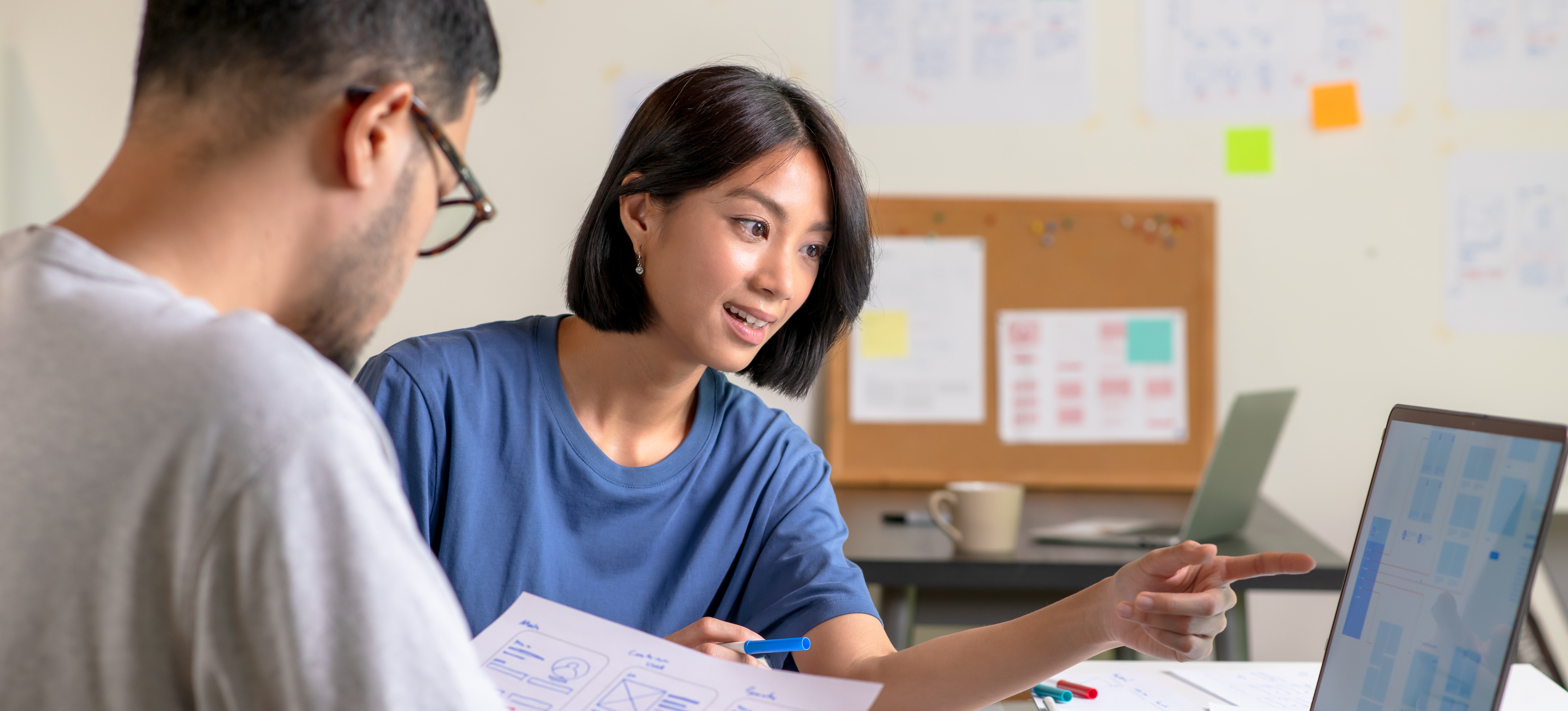 [Featured Image] Two UX designers brainstorming on ideas for a website. They are surrounded by sketches of user flow and design tools. One is gesturing to the laptop that is in front of them, and the other is looking at a paper with sketches.
