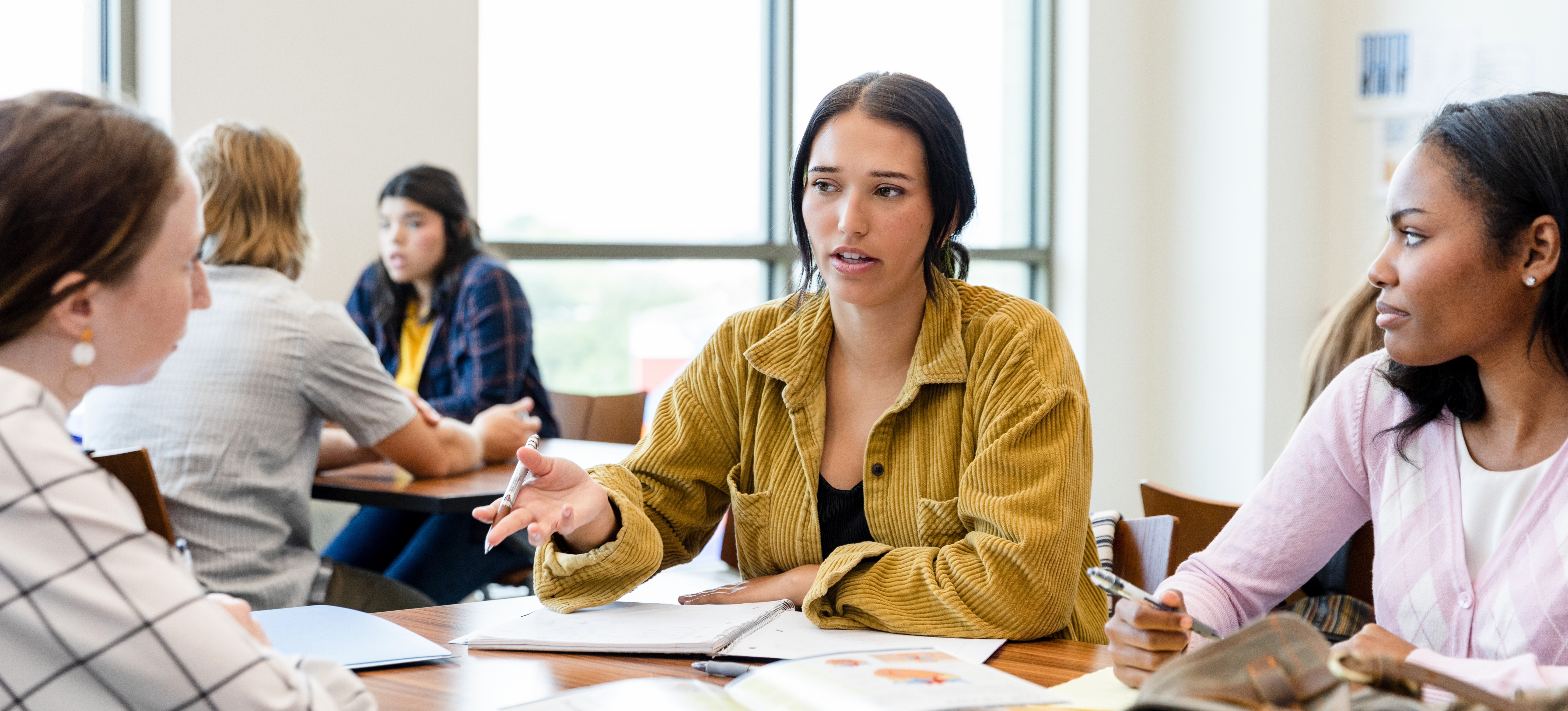 [Featured Image] Three Associate of Arts students study together with books in their college classroom.
