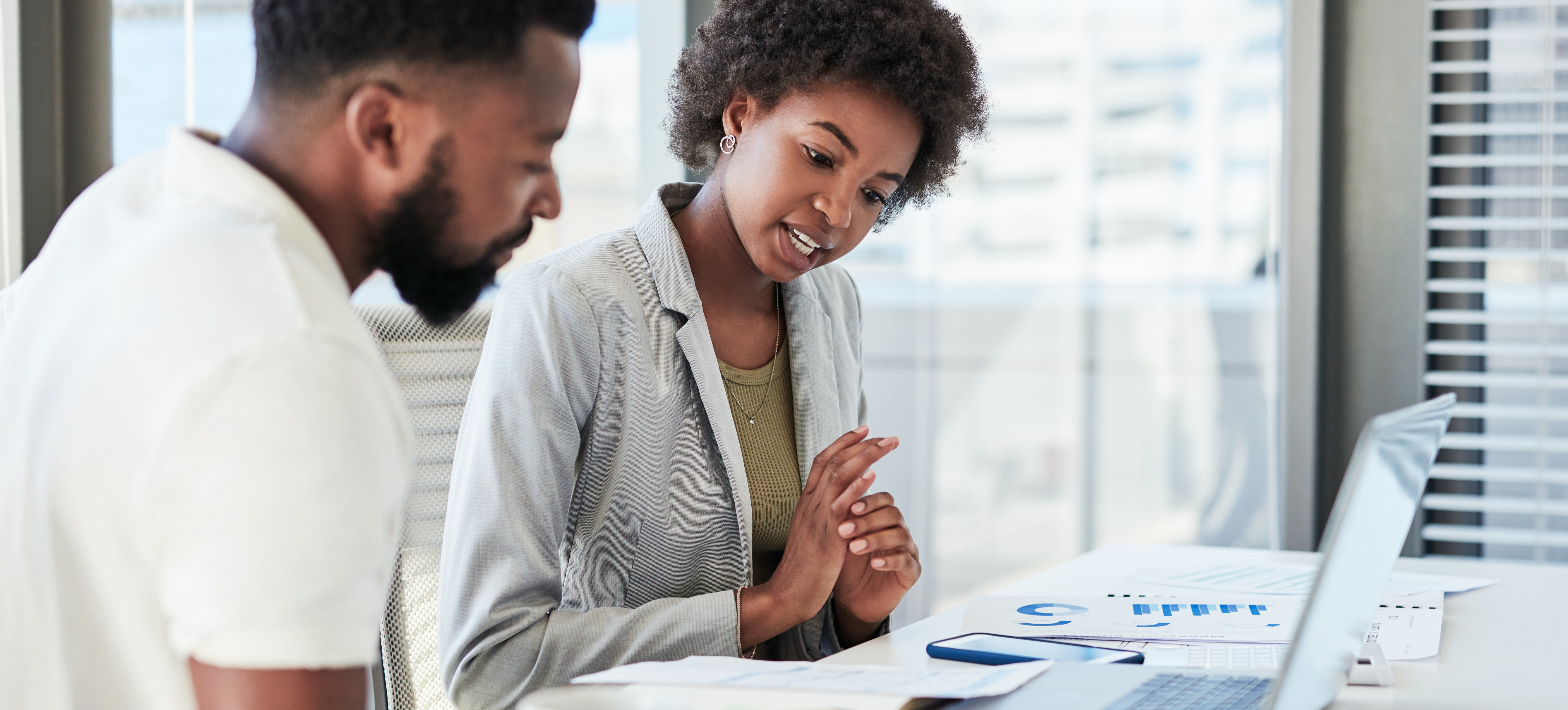 [Featured Image] Two colleagues sit at a desk with a laptop and data they collected through various data collection methods.  
