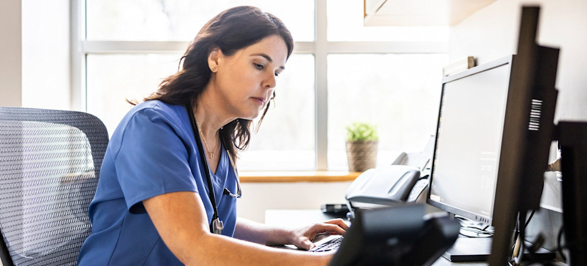 [Featured Image] A nurse is using AI to facilitate their work while adding records to a computer in their office.
