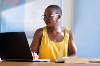 Woman in yellow top, smiling and typing on a laptop.