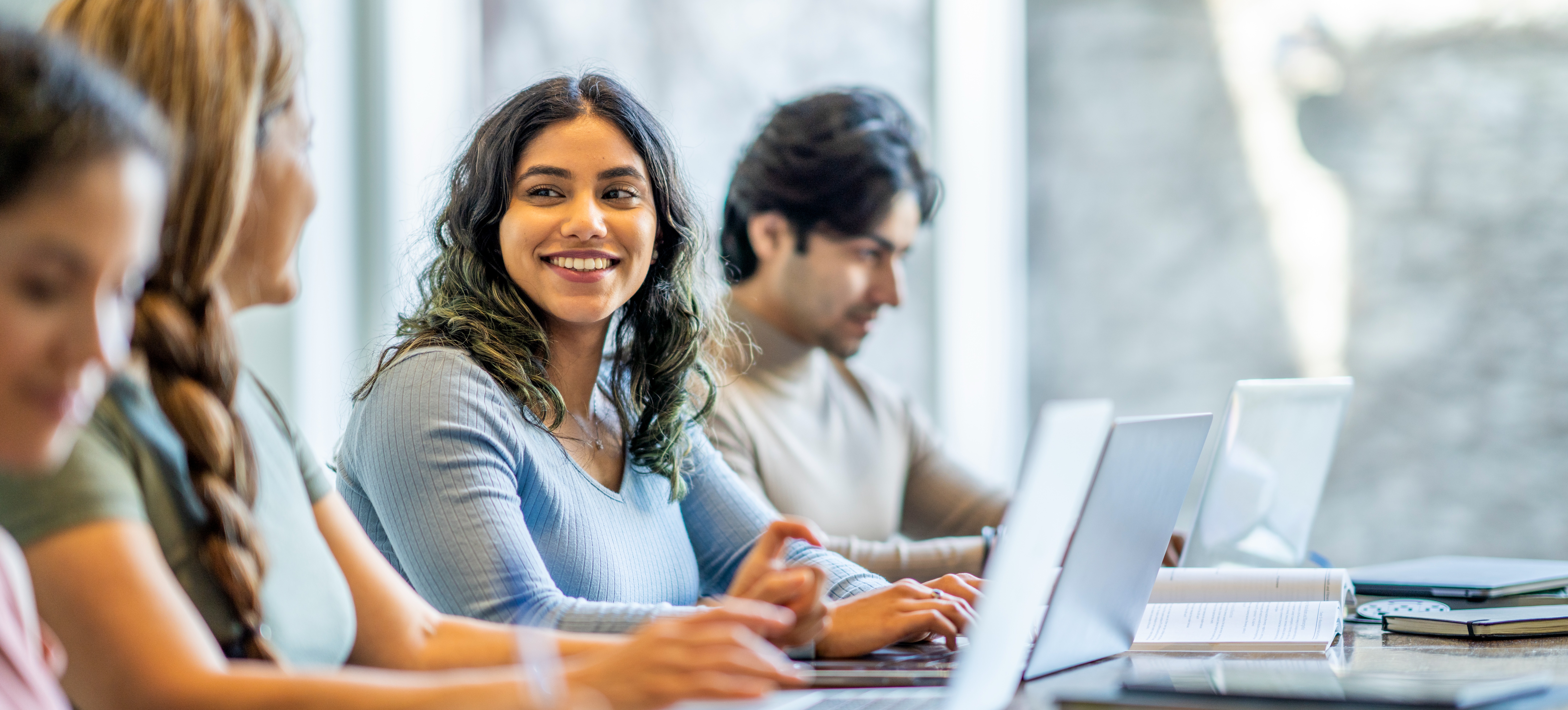 [Featured Image] A doctorate student smiles at their classmate as they sit with other students at a table, working on laptops. 
