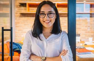 [Featured image] A young graduate student wearing a white button-up shirt and glasses stands with both arms crossed in front of an office. 