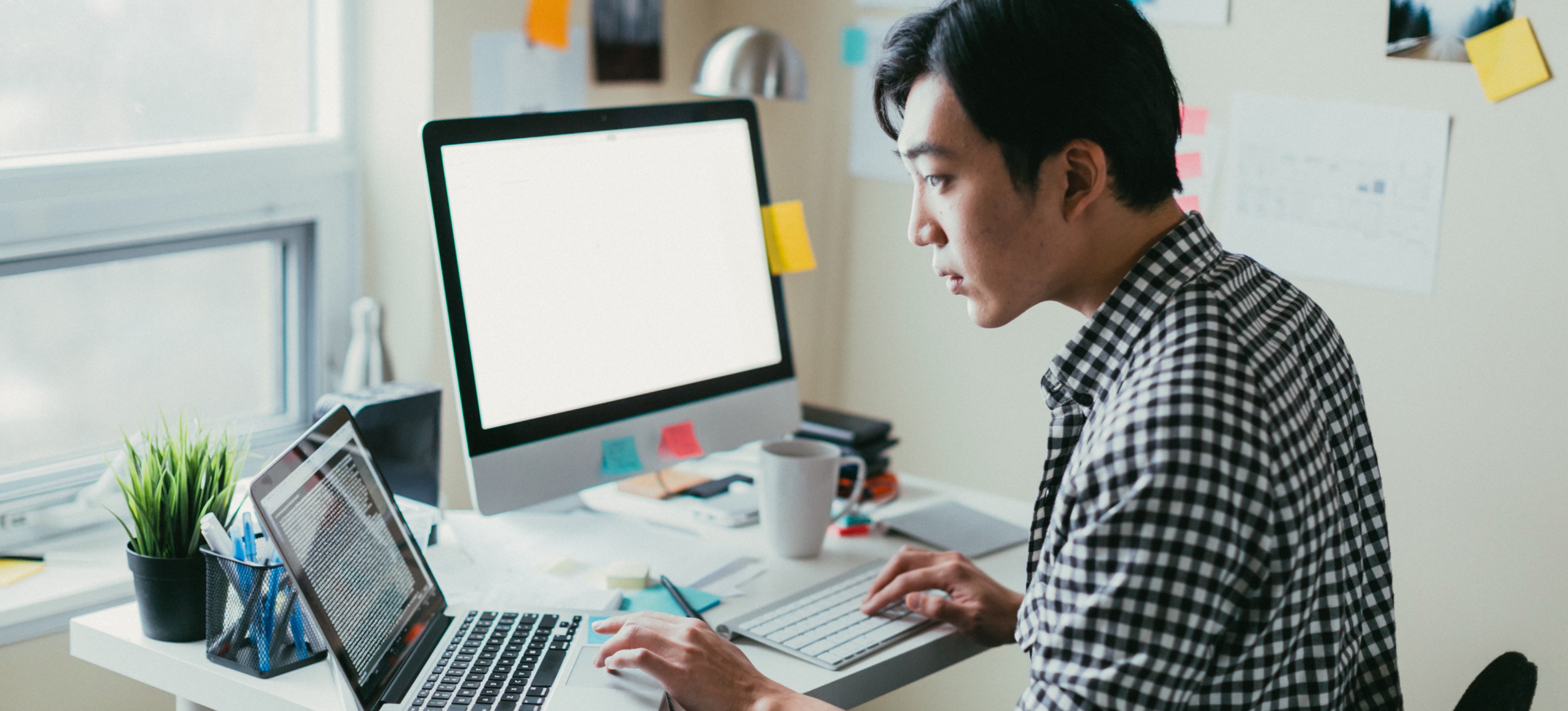 [Hauptbild] Eine Person in einem karierten Button-Down-Hemd arbeitet an einem Schreibtisch mit Laptop, Monitor und Tastatur mit R-Programmierung. Am Monitor und an der Wand rechts hängen farbige Post-Its.