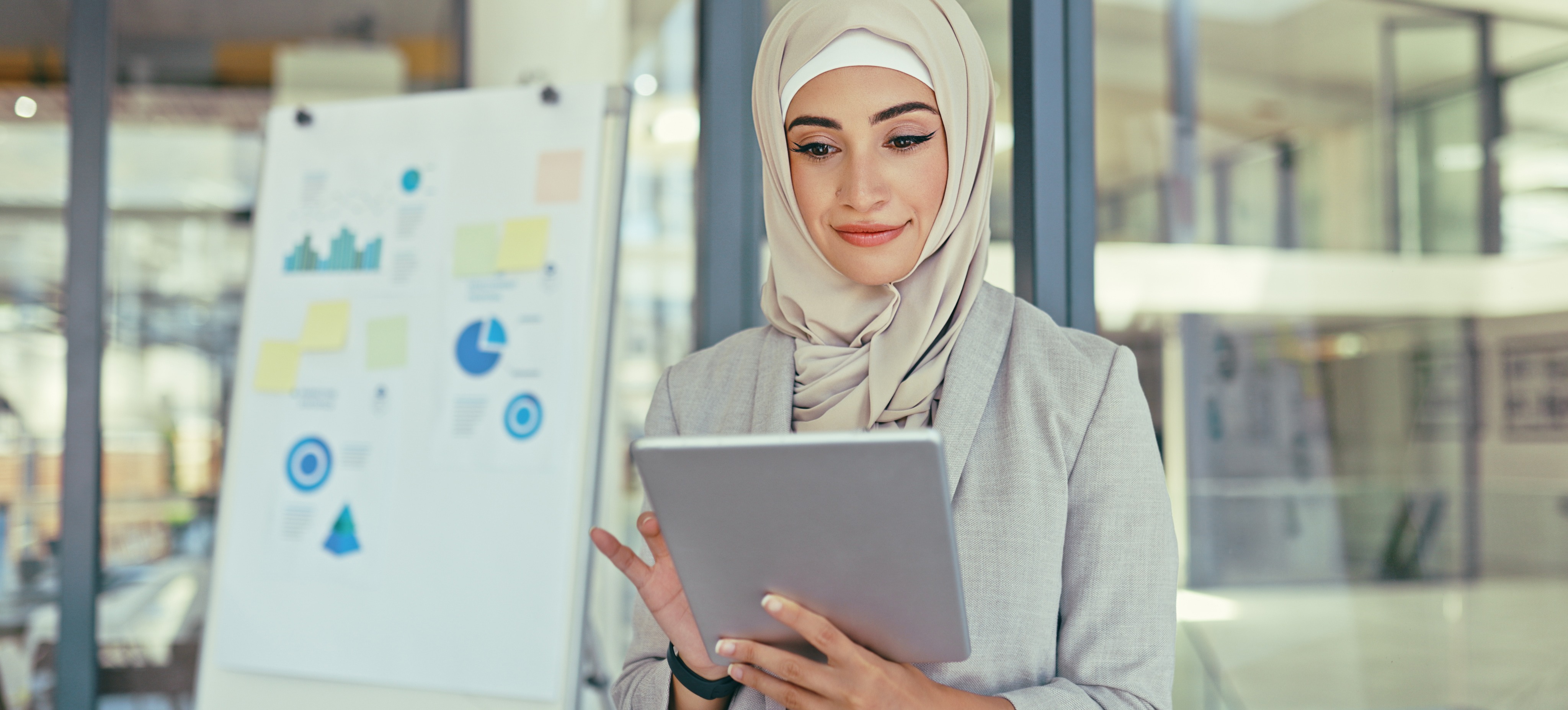[Featured Image] A smiling social media strategist stands in a windowed office looking at their tablet.
