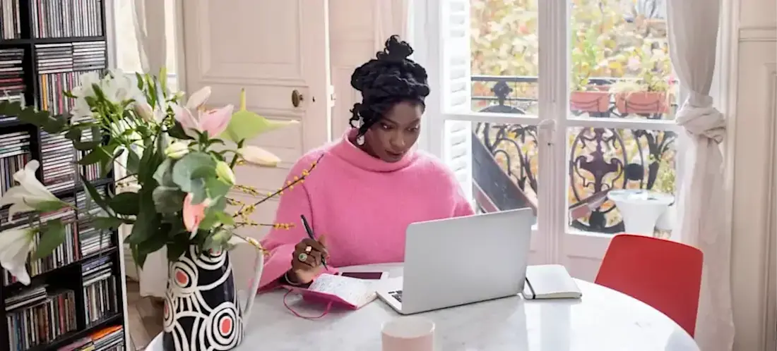 [Featured Image] A person in a pink sweater sits at their dining room table with a laptop in front of a large window, researching which online degree to pursue.