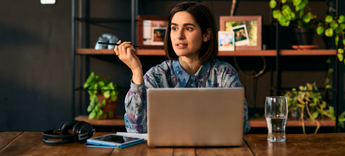 [Featured Image] A home business owner sits at a desk in front of a laptop computer with a pen in their hand, looking off into the distance.
