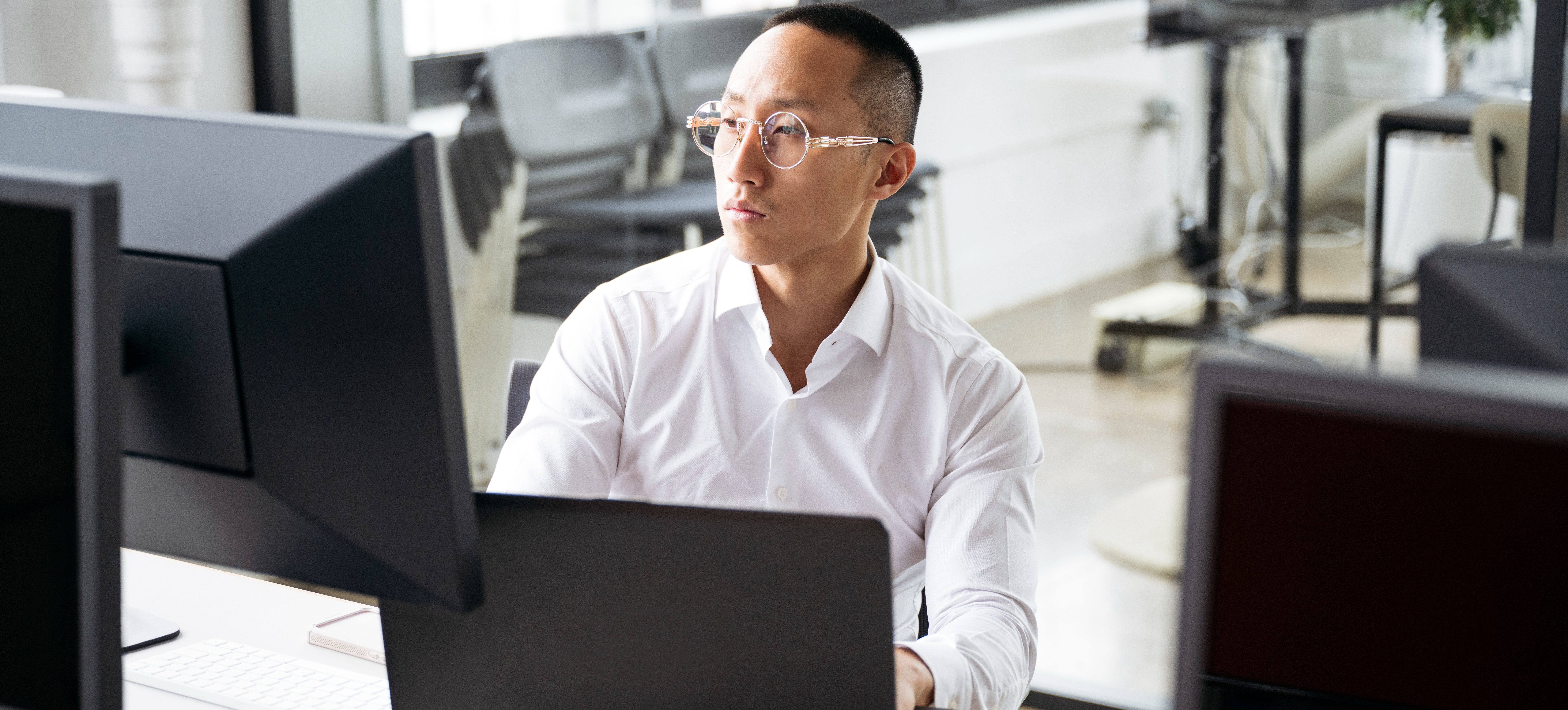 [Featured Image] A data analyst sits at his office computer and laptop, working on business intelligence analytics.