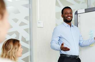 [Featured Image] A project manager in a blue shirt stands in front of a whiteboard and presents to three other people in a conference room.