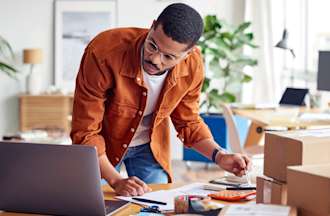 [Featured image] Dark skinned man with round glasses packs boxes for his ecommerce business from home and leans over table doing calculations on the calculator.