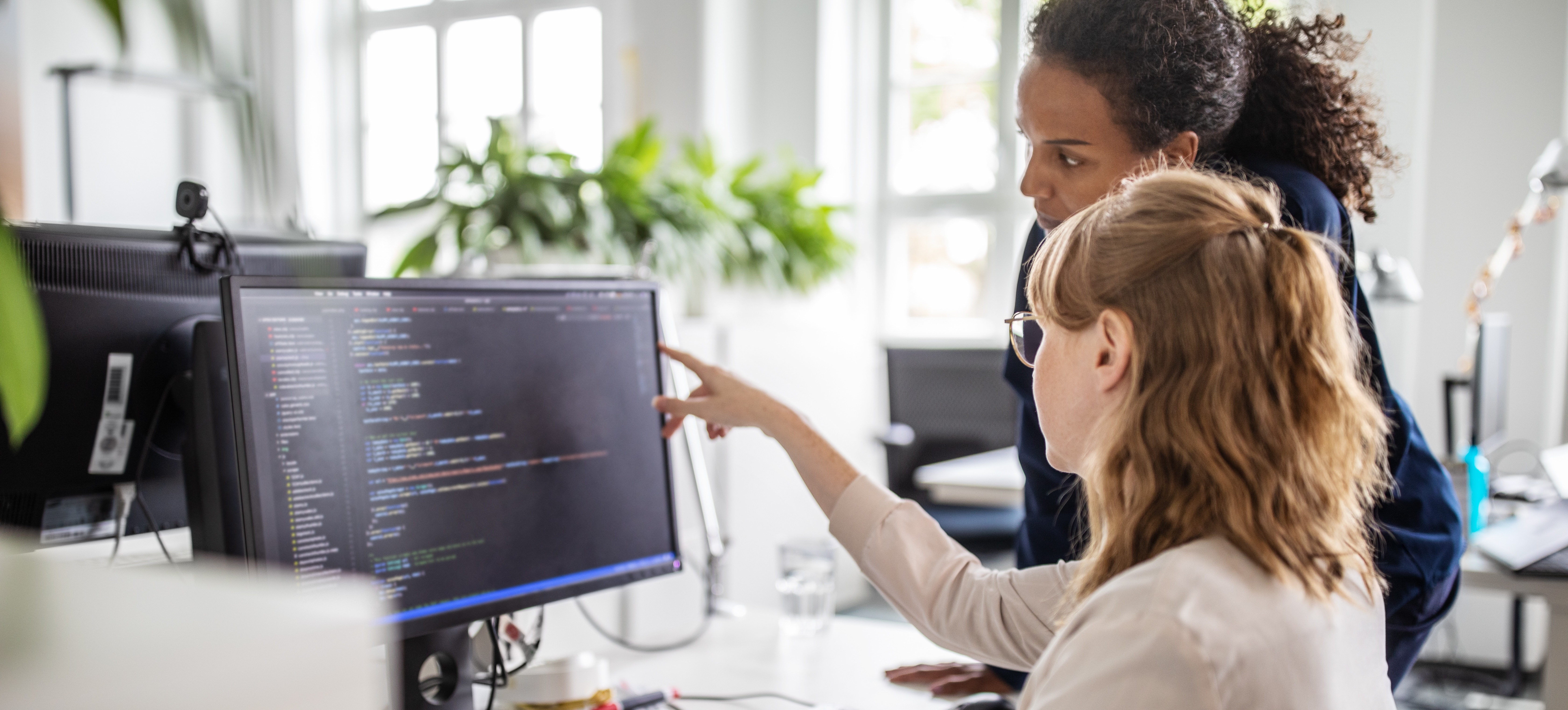 [Featured Image] In an office, a woman points at a computer screen filled with code, as she and a woman colleague discuss software integration.
