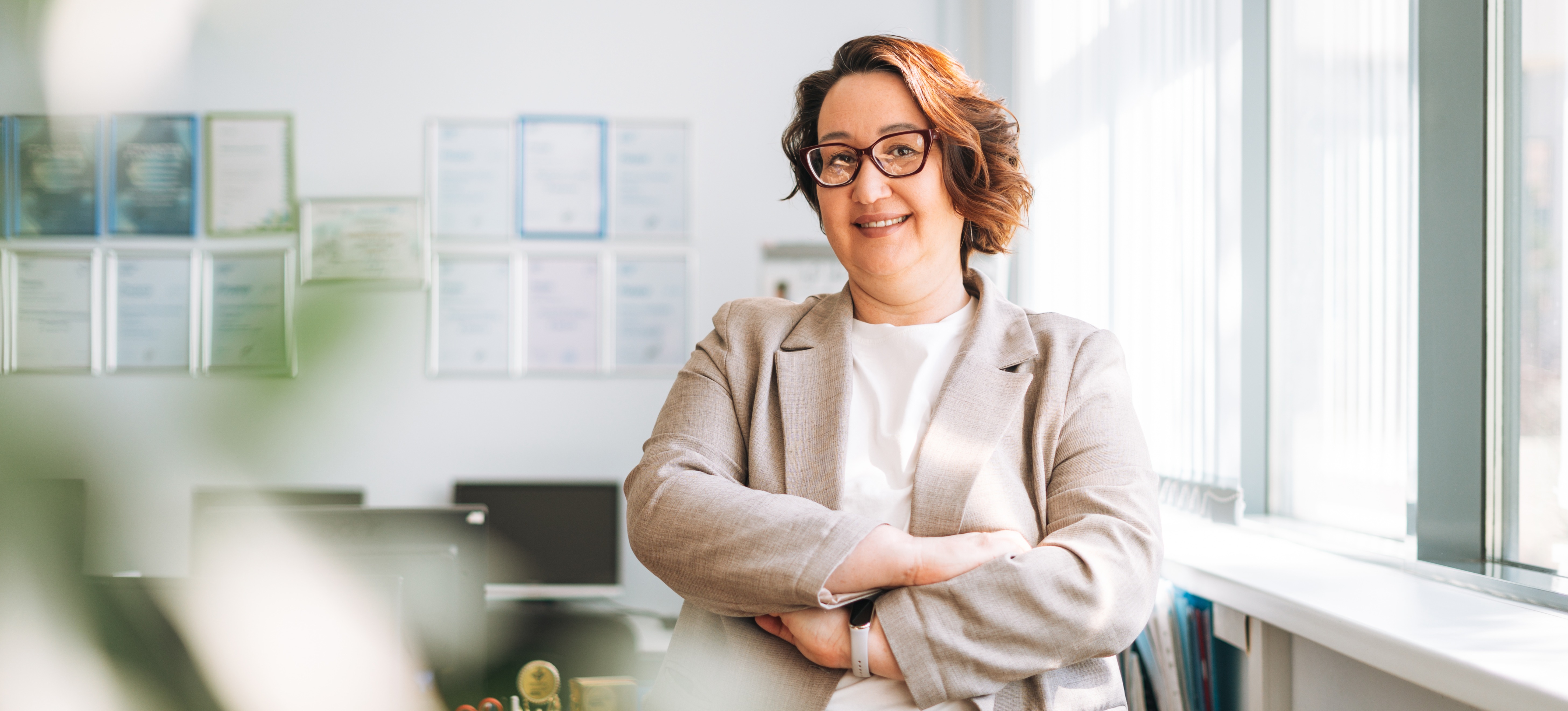 [Featured image] An accountant in a beige blazer and glasses stands by their desk with their arms crossed while looking at the camera and smiling.
