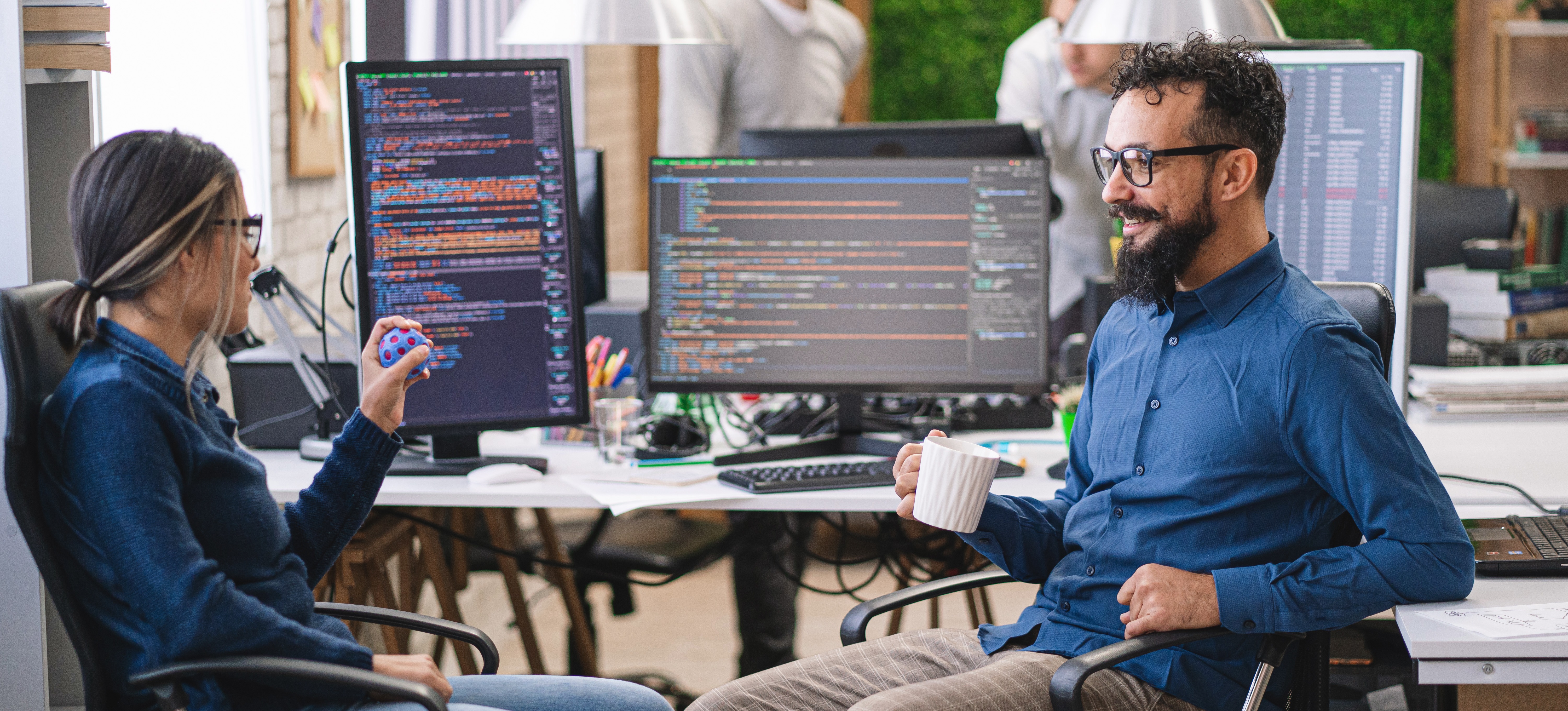 [Featured Image] Two members of a DevOps team discuss continuous integration systems while sitting at their desks with several computer monitors, and two additional team members are working in the background.
