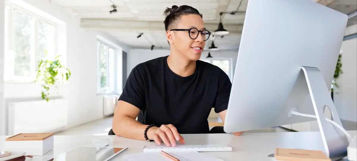 [Featured Image] A person in a black shirt works in an office at a desktop computer. 