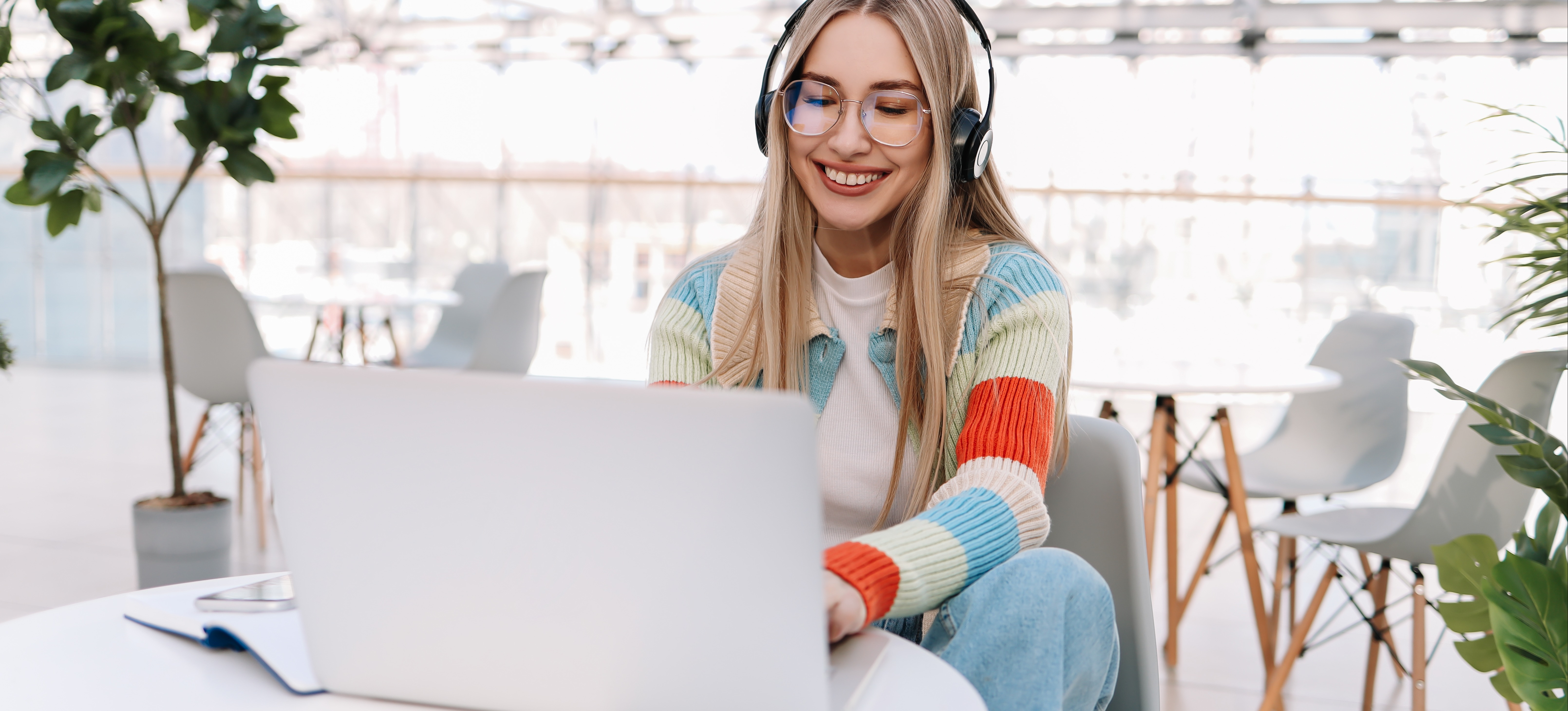 [Feature Image] A prospective learner listens to music in headphones while researching business degrees FAQ on their laptop in a sunny atrium.
