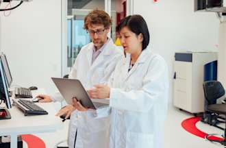 [Featured image] A physician assistant in a white lab coat shows test results on a laptop to her colleague.