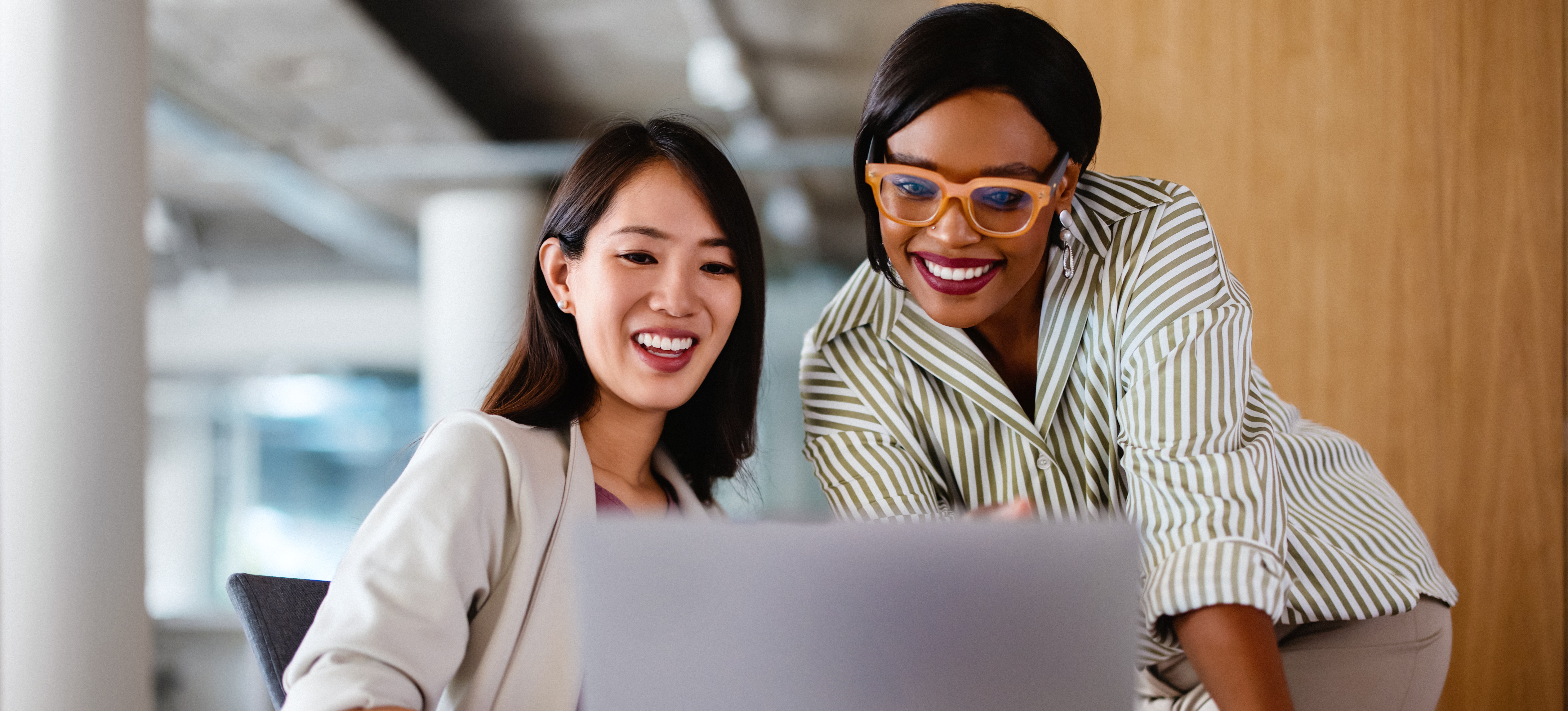 [Featured Image] Two professional women sit at a computer and discuss what an operating system is and which one they prefer. 