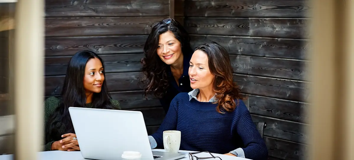 [Featured image] A business person with brown hair and blue sweater chats with two colleagues about a proposal on their laptop.