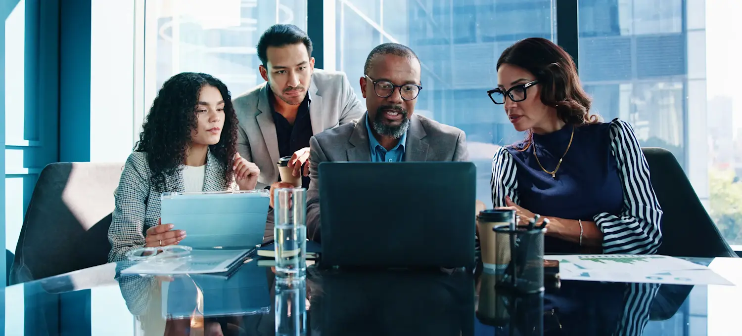 [Featured Image] A business team seated at a glass conference table in front of a large office window discusses AI risk management as they look at a laptop screen.

