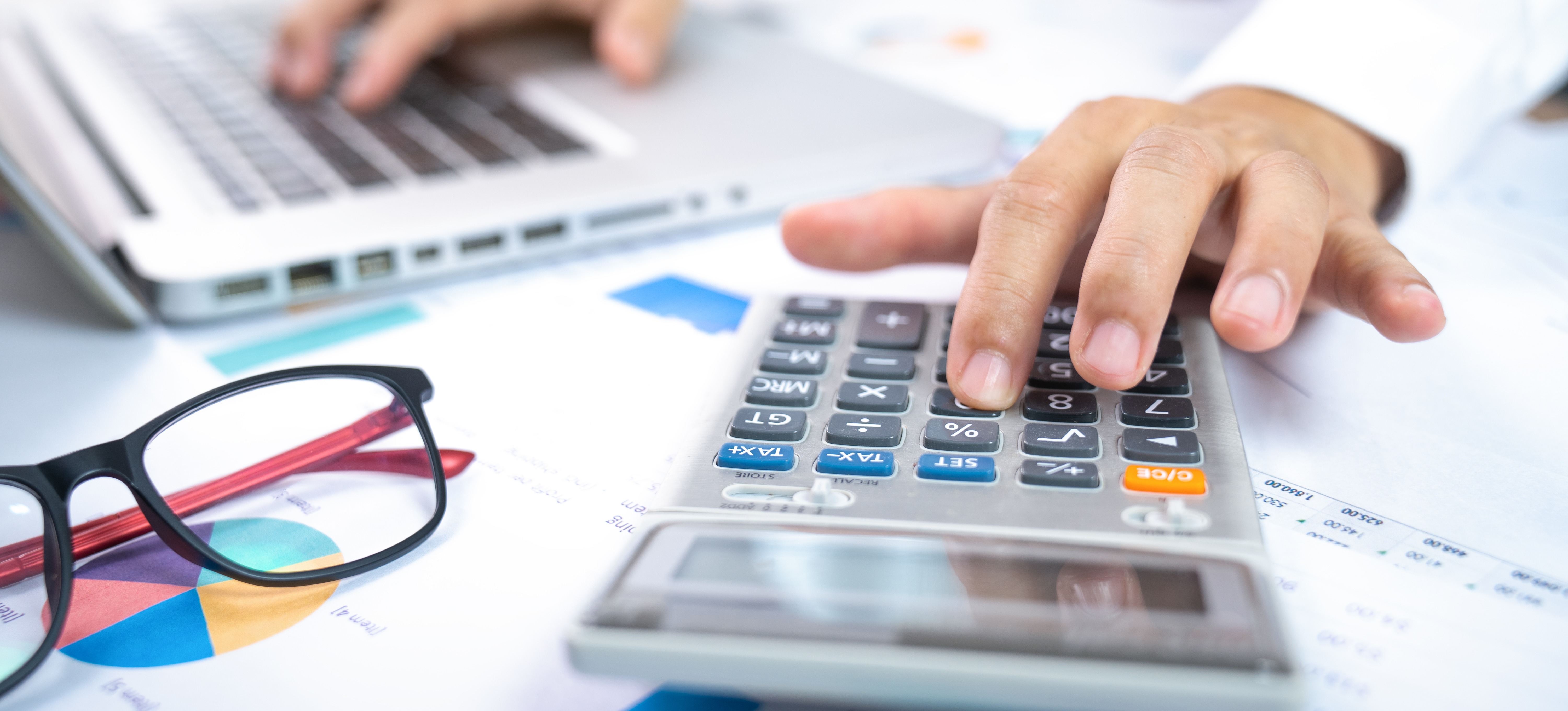 [Featured Image] A person's hand is shown in the foreground touching the buttons of a calculator resting on papers with graphs on them while the other hand in the background types on a computer keyboard. 