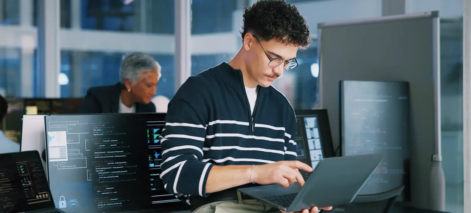 [Featured Image] A web developer goes over SSL/TLS protocols as they sit on the edge of their desk in a shared office and work on a laptop.
