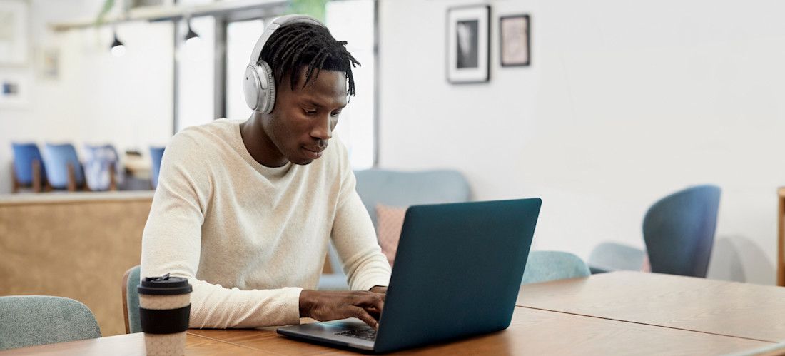 [Featured Image] A PhD student works on his laptop while listening to over-the-ear headphones. 