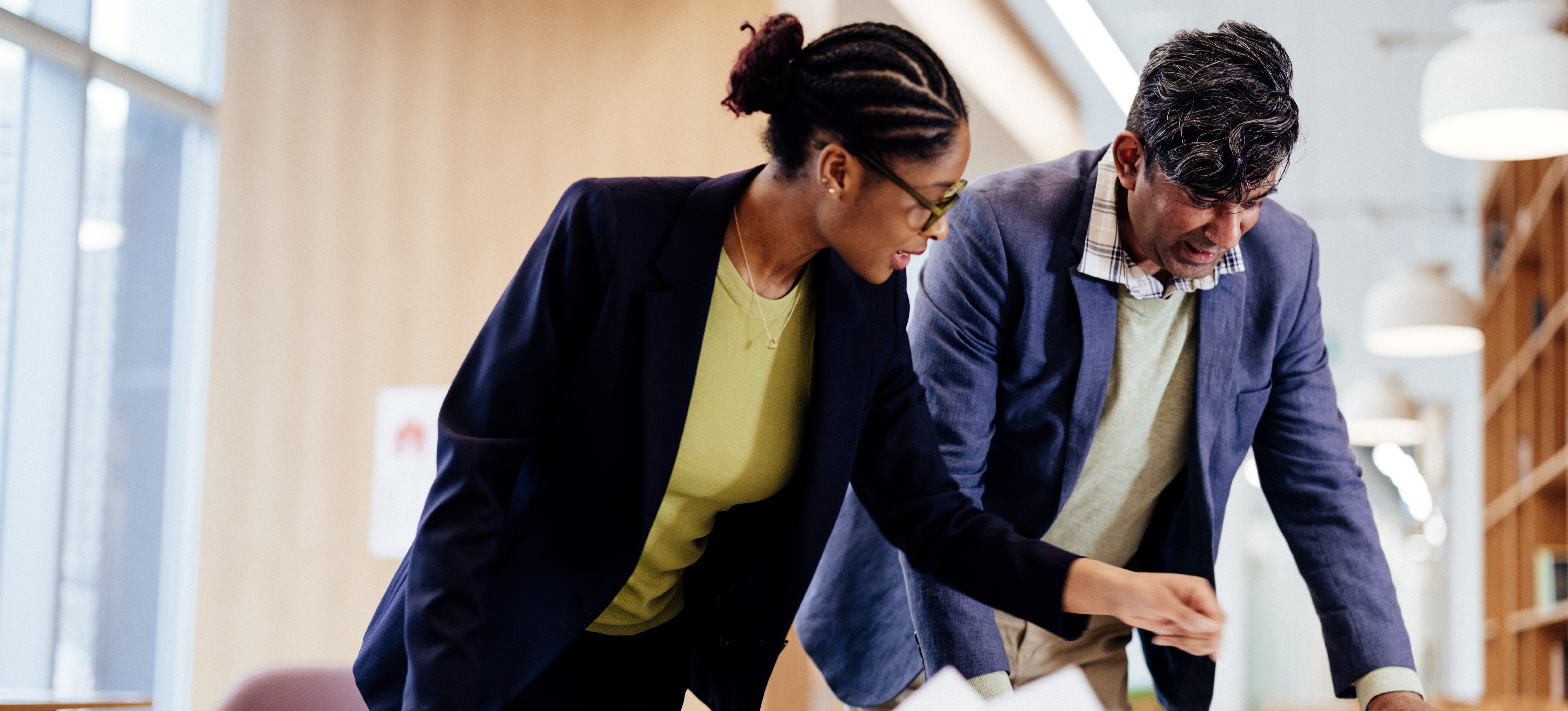 [Featured Image] A project coordinator works a high-paying job without a degree in a conference room, showing blueprints on a table to a coworker.
