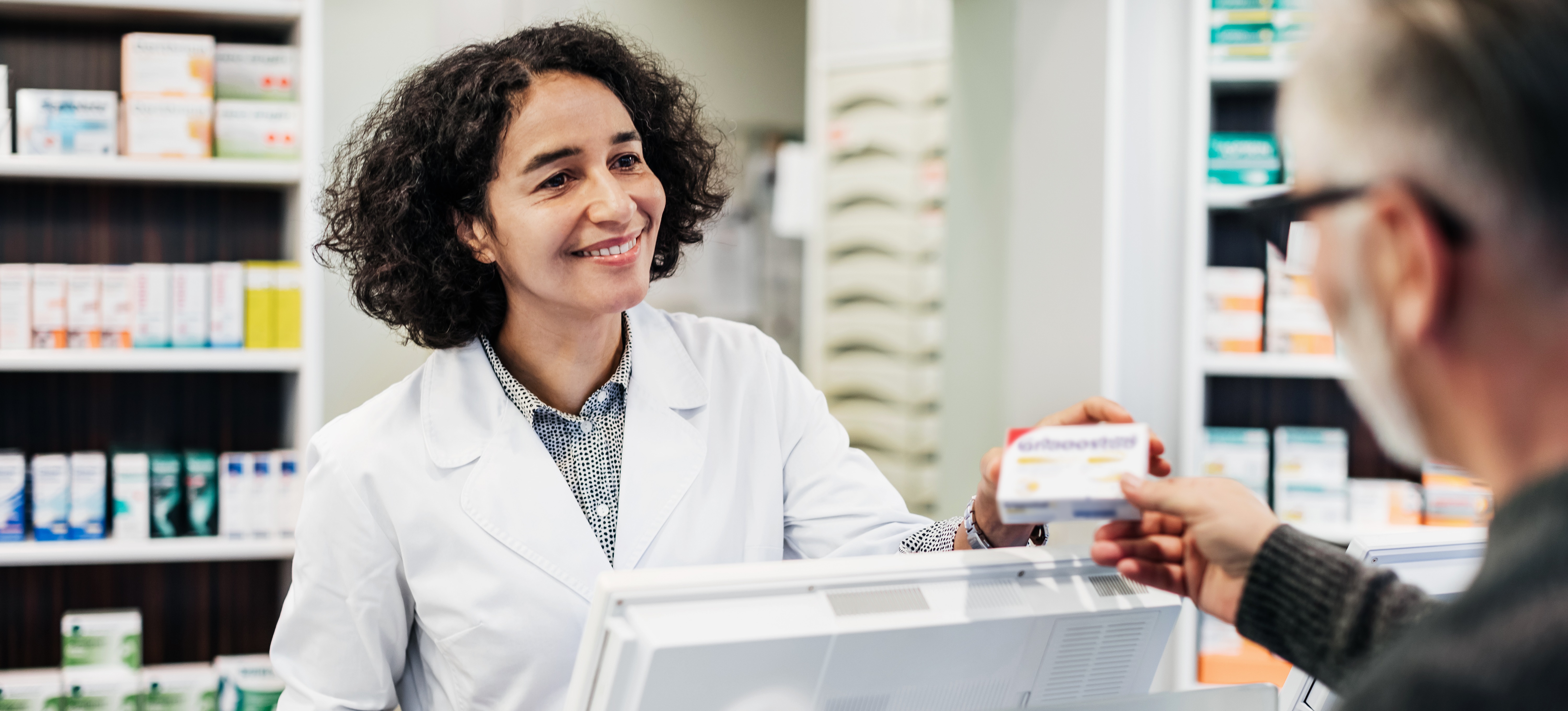 [Featured Image] A pharmacist smiles while handing a prescription to a customer.
