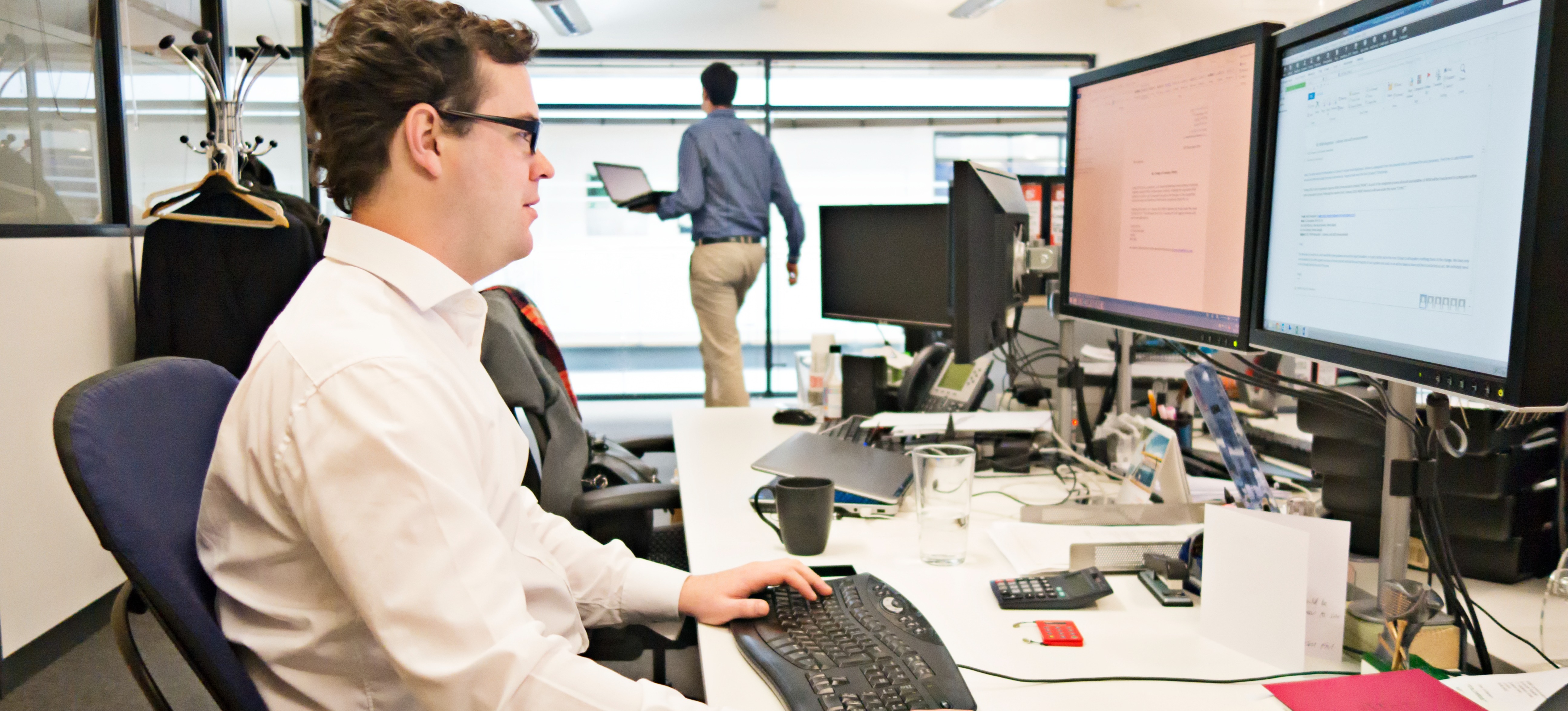 [Featured Image] A technology consultant works at an office desk using a computer with two monitors as colleagues walk in the background.
