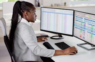 [Featured Image] A data analyst sits at her office desk analyzing spreadsheets on her two computer screens to gain data insights about her work.
