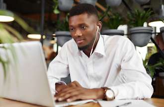 [Featured Image] A prompt engineer in a white button-down shirt sits at a table and works on their laptop. There is a pad and pen to their left and a wall of plants behind them.