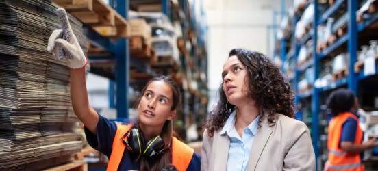 [Featured image] Two product managers are wearing an orange vest, and gloves looking at warehouse products. 