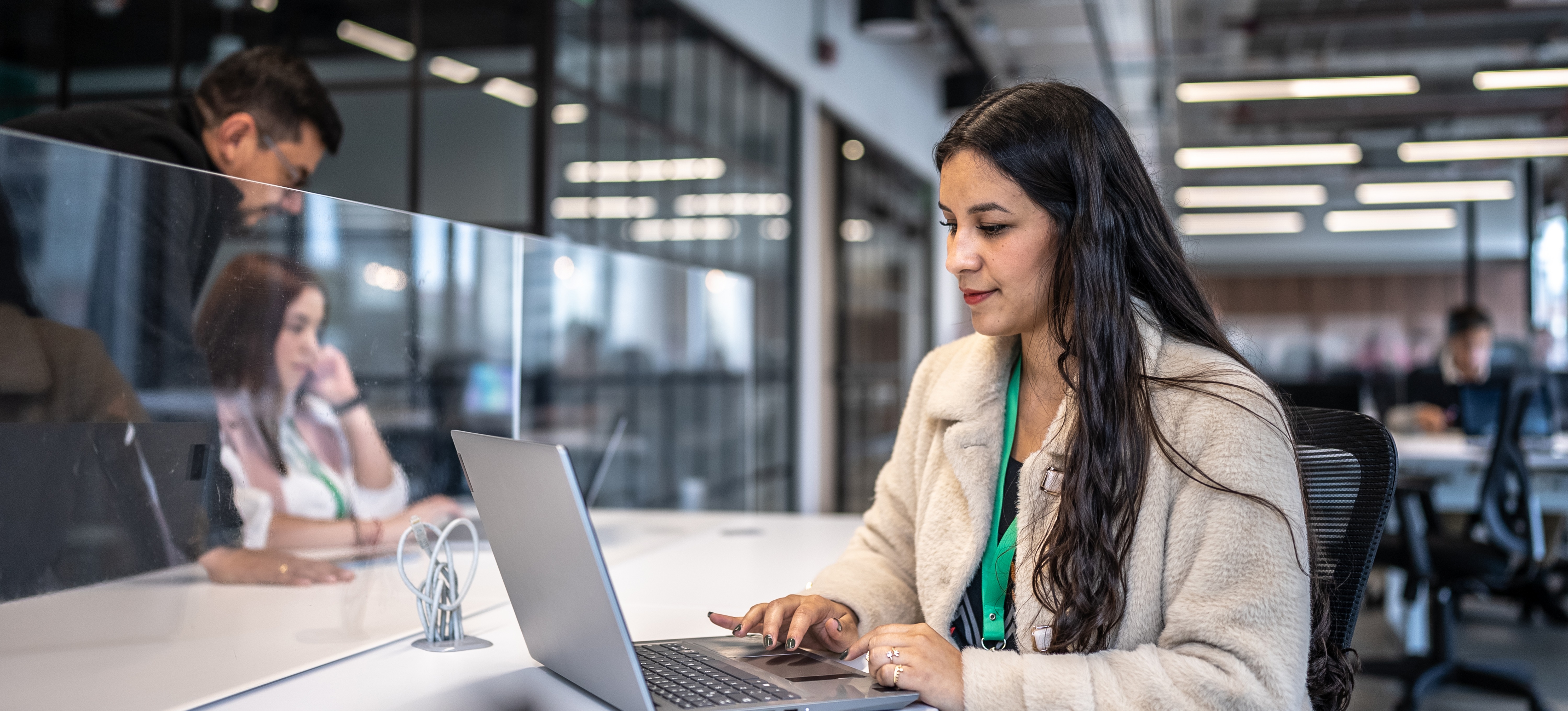 [Featured Image] An InfoSec professional with long black hair, a beige jacket, and an ID badge sits at her laptop in an office environment while her colleagues work at surrounding work stations.
