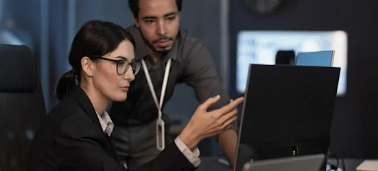 [Featured image] A cybersecurity analyst consults with a colleague while pointing at a computer monitor.