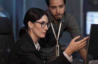 [Featured image] A cybersecurity analyst consults with a colleague while pointing at a computer monitor.