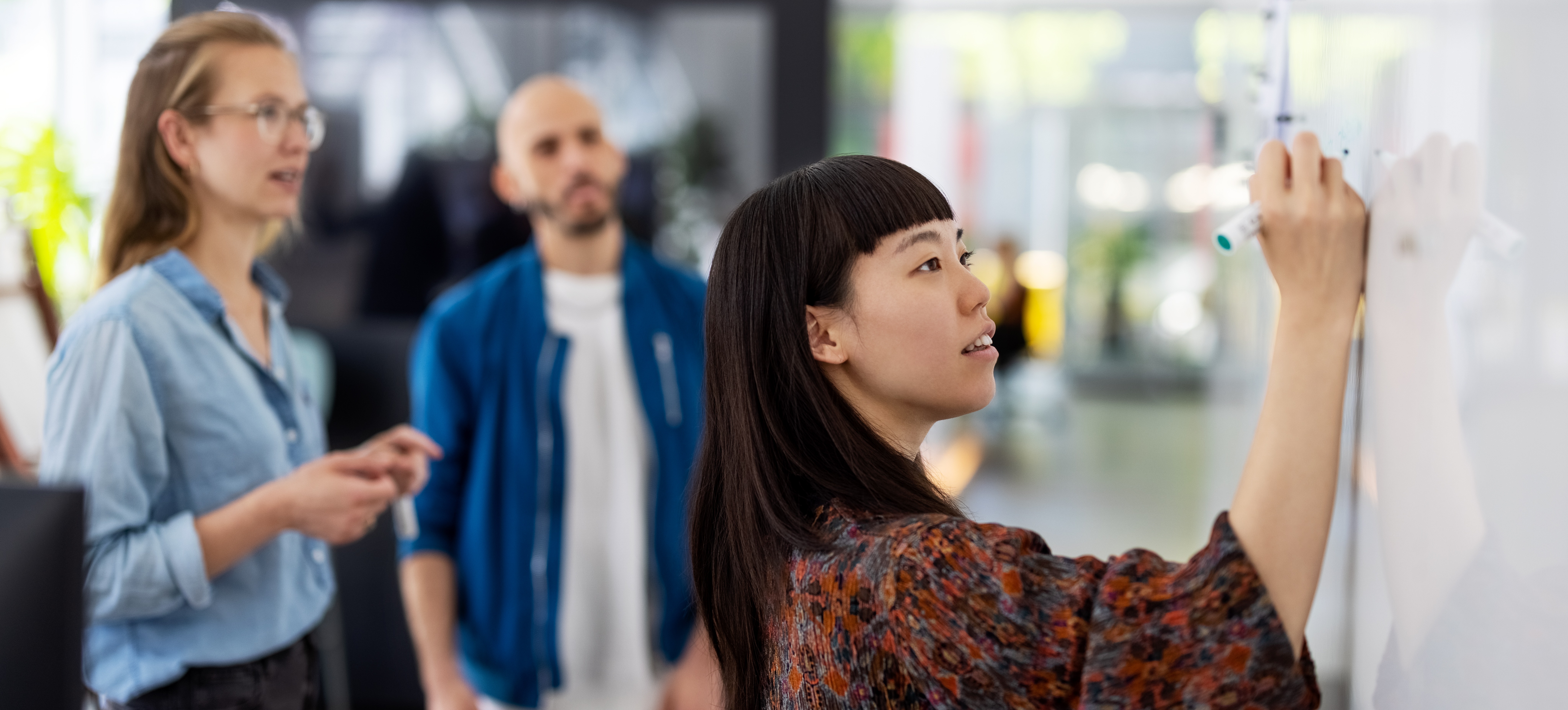 [Hauptbild] Ein Projektmanager verfolgt mit zwei Kollegen Projektmeilensteine ​​auf einem Whiteboard.
