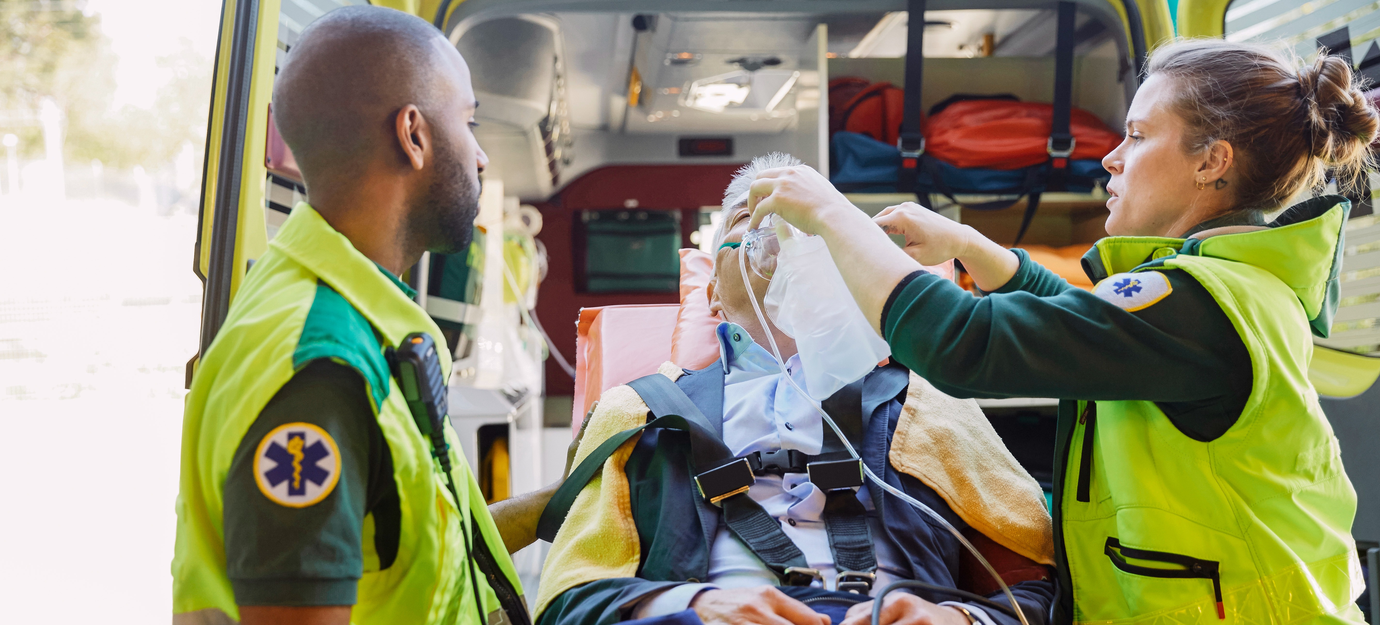 [Featured Image] Two EMTs with emergency management degrees dressed in bright yellow uniforms stand outside of an ambulance and attend to a patient in a stretcher.
