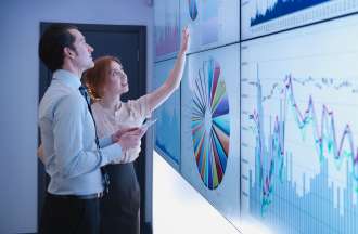 [Featured Image] Two co-workers review performance data in a set of visualizations displayed across a series of screens on a wall.