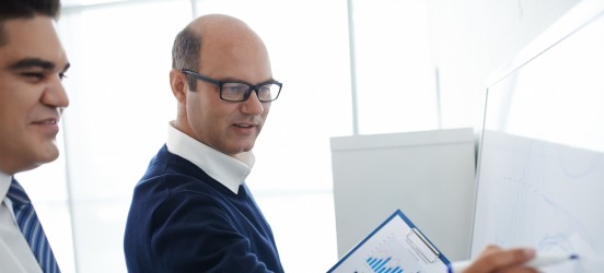 [Featured Image] A project manager is standing at a board and holding documents with graphs and tables, discussing the latest project strategy with his team member.