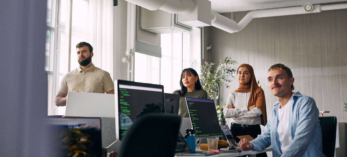 [Featured Image] A DevOps team listens to a team member as they sit and stand at a workstation with computers.
