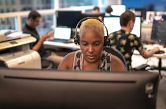 [Featured image] An information security analyst wearing black headphones and a patterned tank top works at a monitor with a printer to their right and colleagues in the background.