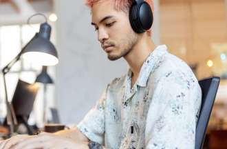 [Featured Image] Side view of a young IT professional wearing a headset, typing at a laptop, and using a second computer monitor while coding at a coworking office space.
